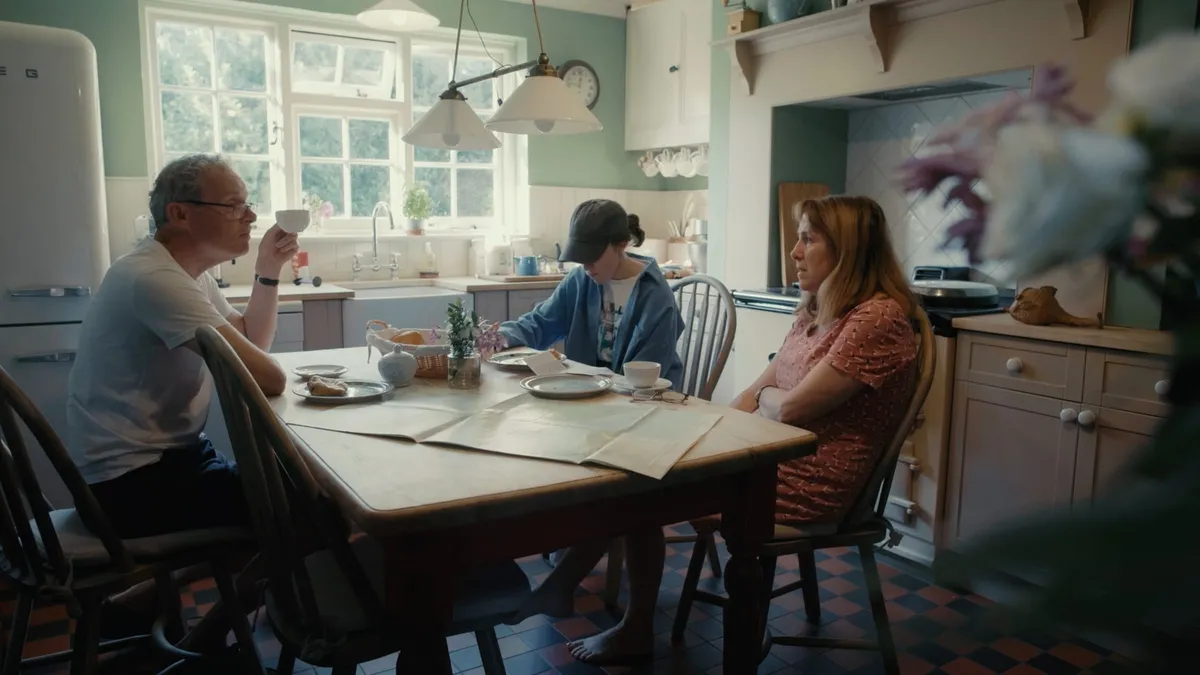 A grey-haired man in glasses and a white T-shirt drinks from a cup at a kitchen table, a boy in a cap and a woman in pink seated with him, papers spread on the table.
