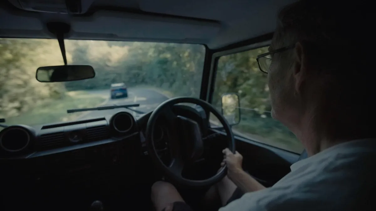 Interior view from behind a grey-haired man in glasses driving a vehicle along a tree-lined road, another car ahead through the windscreen.