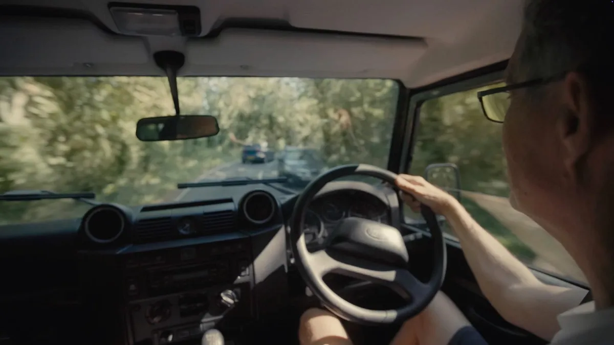 Interior view from behind a grey-haired man in glasses driving a vehicle along a tree-lined road, a car ahead through the windscreen.