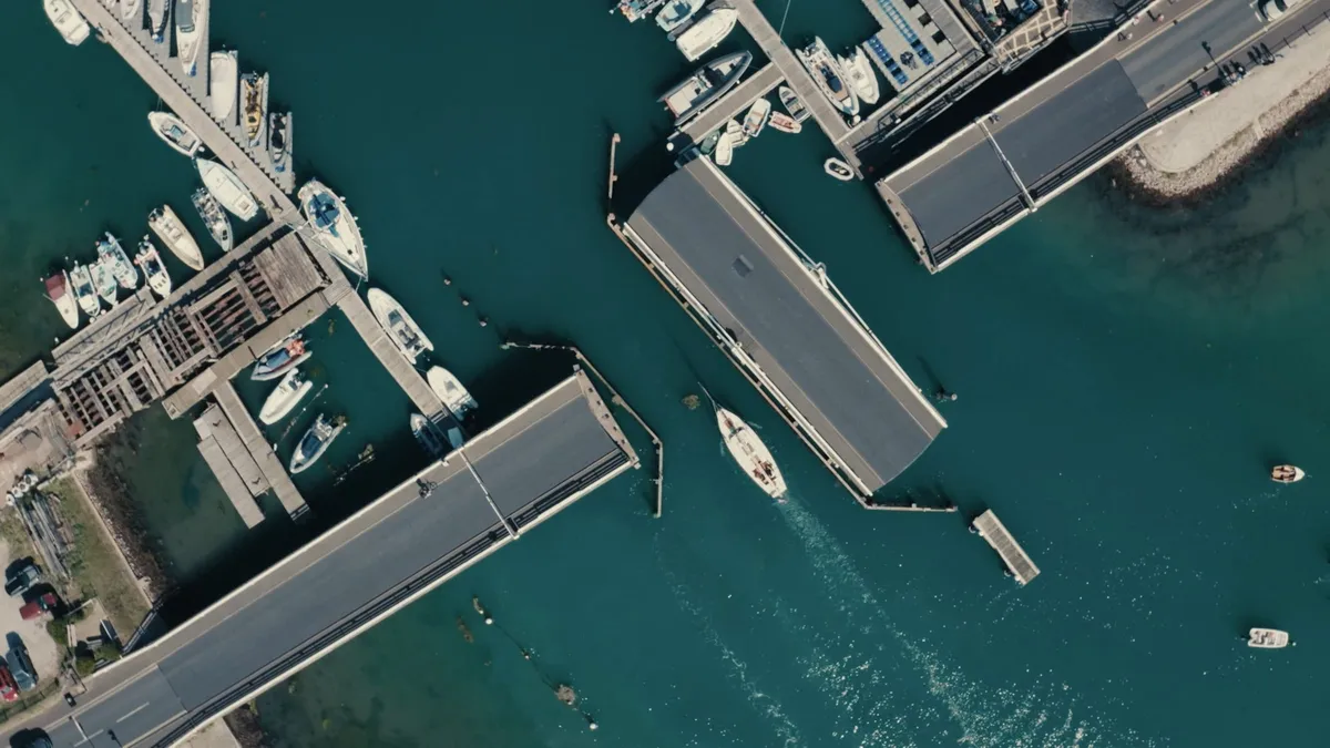 Aerial view of a marina with two swing bridges open over a channel, a yacht passing between them and boats moored along wooden jetties.