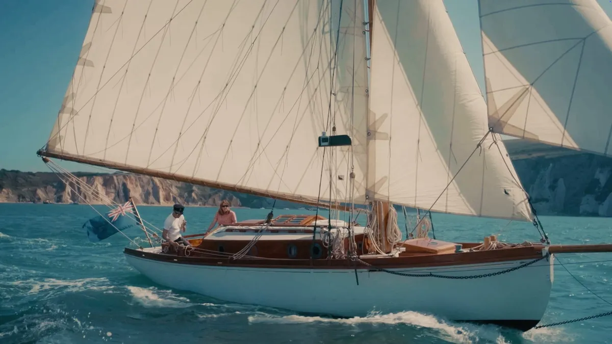 A white sailing yacht with full sails passes chalk cliffs, two people at the helm, sea spray at the bow.