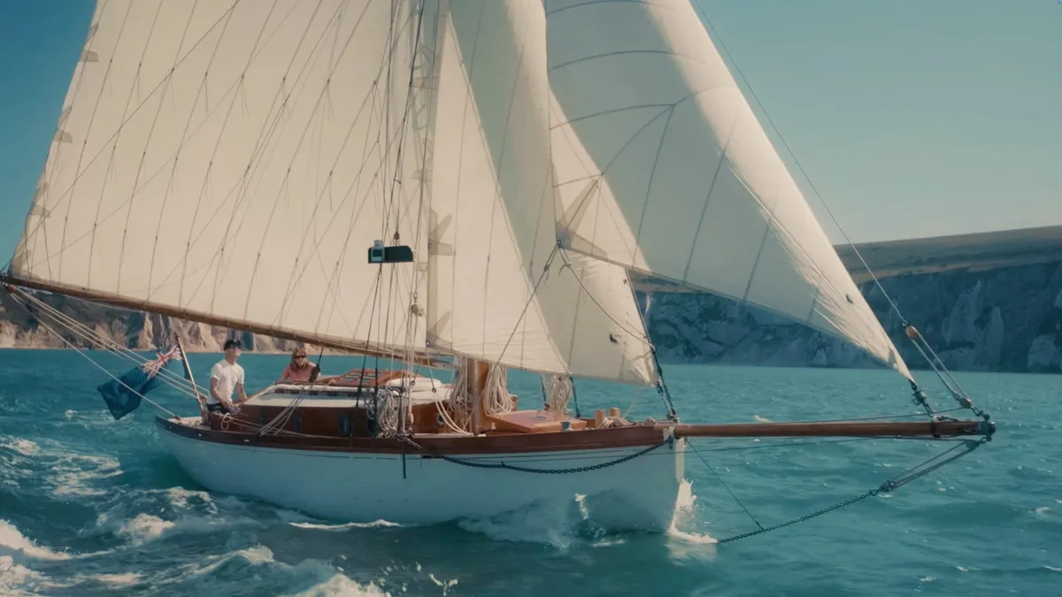 A white sailing yacht with full sails passes chalk cliffs, two people at the helm, wake trailing behind in blue water.