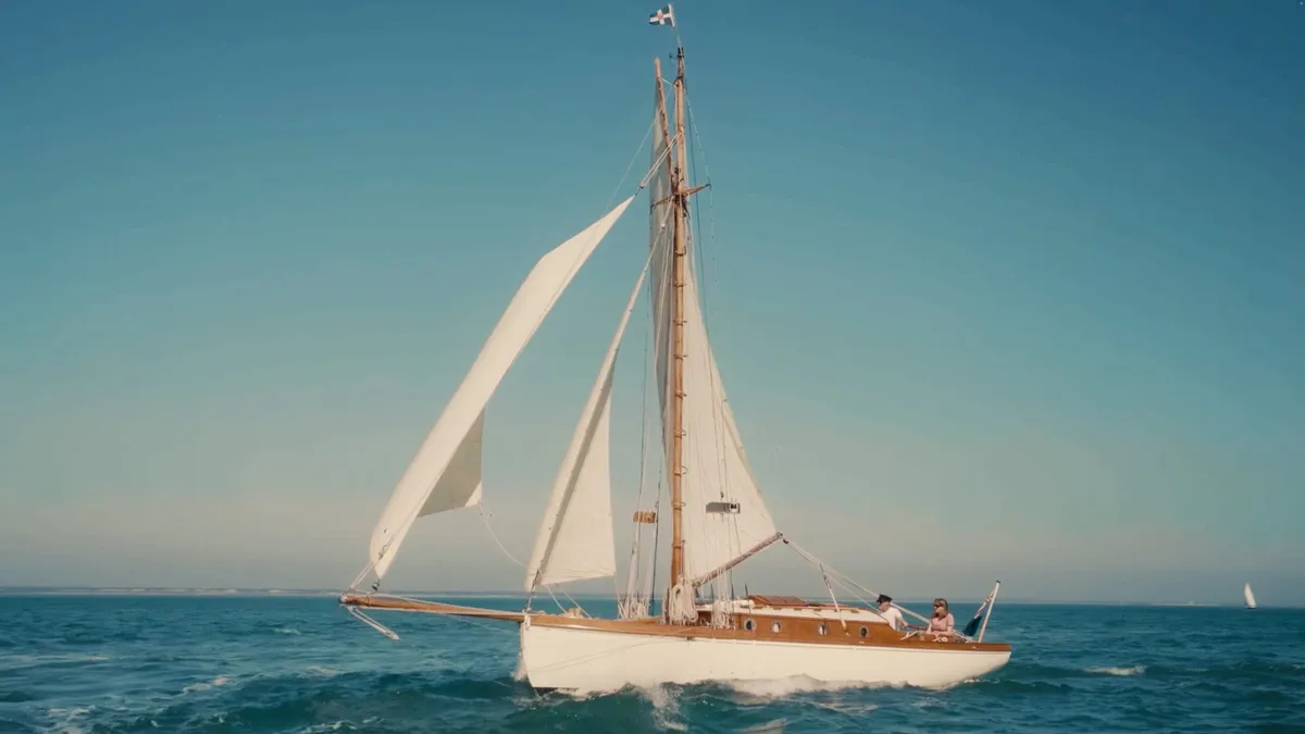 A wooden sailing yacht with three raised white sails heels across open water, two people seated at the helm, a distant sail visible on the horizon.