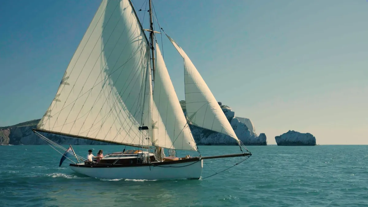A white sailing yacht with three raised sails passes chalk sea stacks, two people seated at the helm, calm blue water around the hull.
