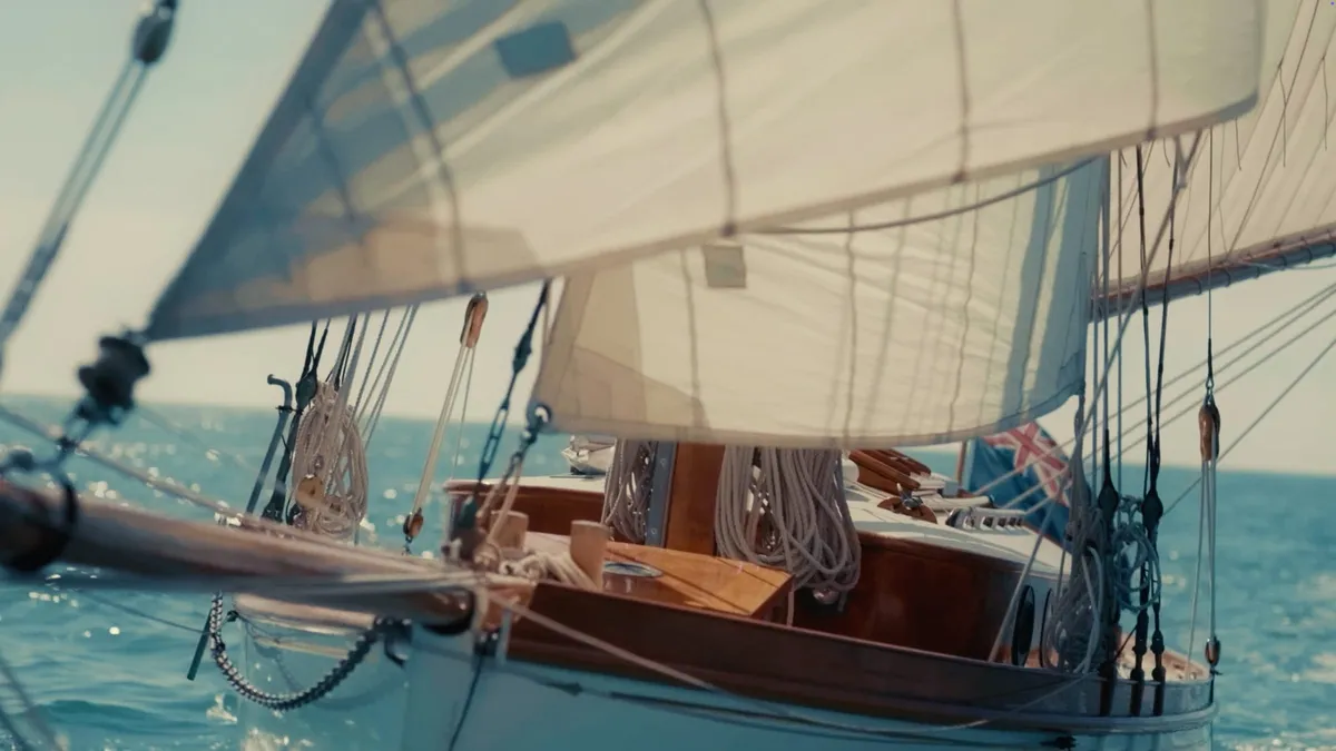 Close-up of a wooden sailing yacht's deck and rigging, sails billowing overhead, coiled ropes on deck and a flag at the stern.