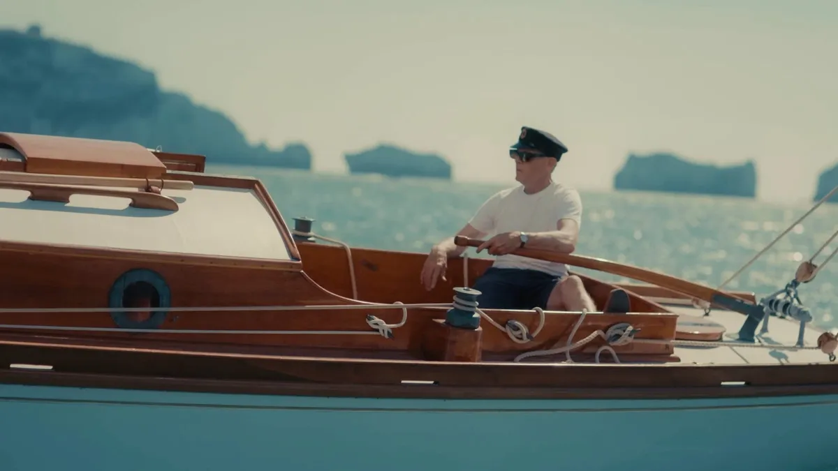 A man in a peaked cap and sunglasses, wearing a white T-shirt and dark shorts, sits steering a wooden boat, sea stacks visible behind him.