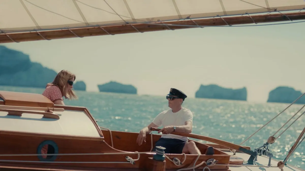 A man in a peaked cap and sunglasses, wearing a white T-shirt and dark shorts, steers a wooden sailing boat while a blonde woman in a pink top leans in behind him, sea stacks in the distance.