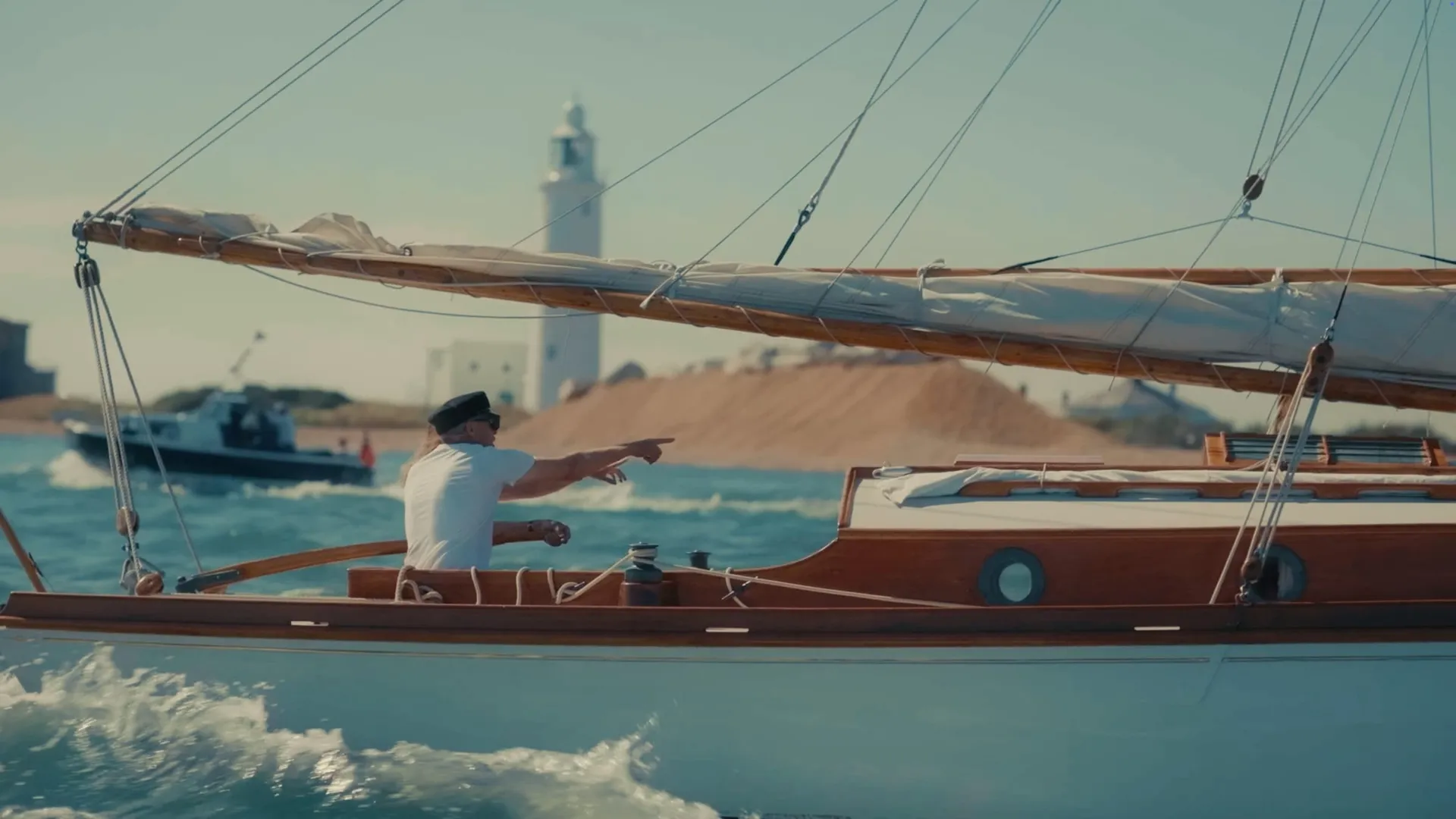 A man in a peaked cap and white T-shirt points from a wooden sailing boat, a lighthouse and small boat visible behind him on the water.