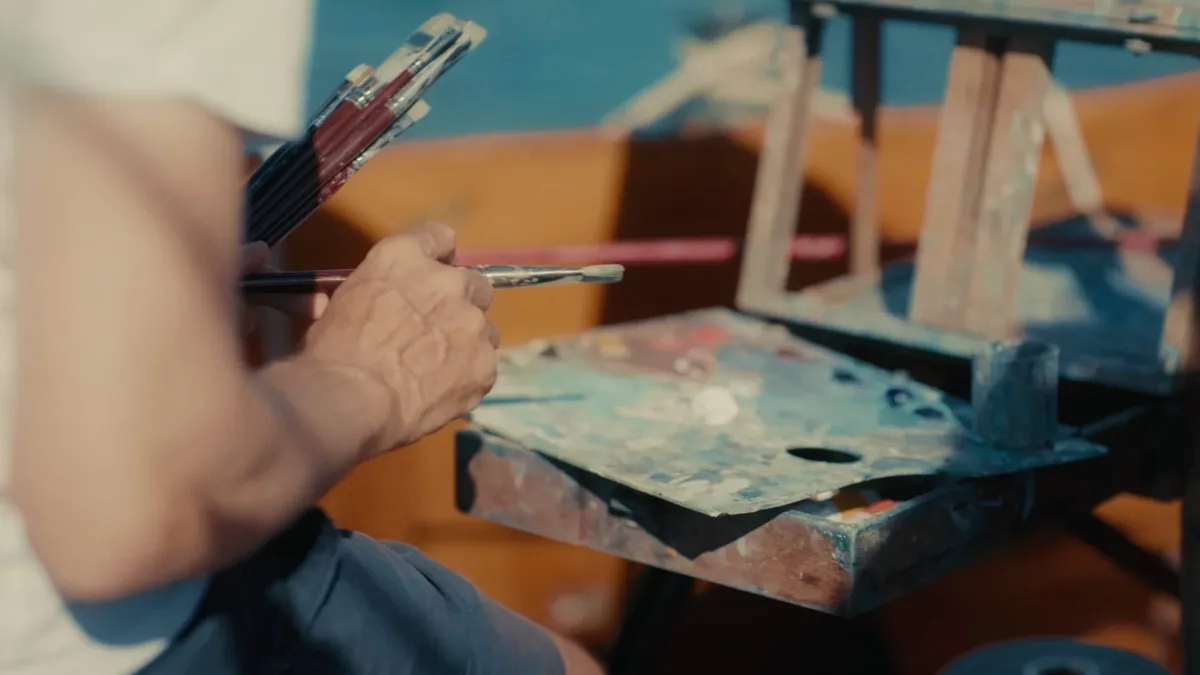 Close-up of a hand holding paintbrushes beside a paint-loaded palette on an easel, a boat's mast and orange hull blurred behind.