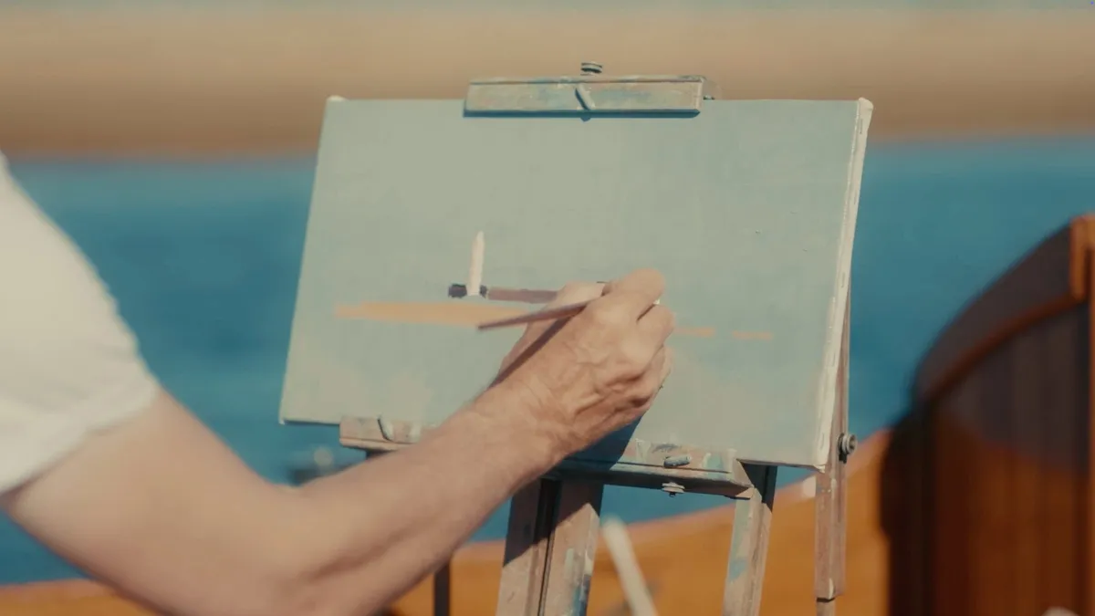 Close-up of a hand painting a small boat onto a pale blue canvas on an easel, sea visible in the background.