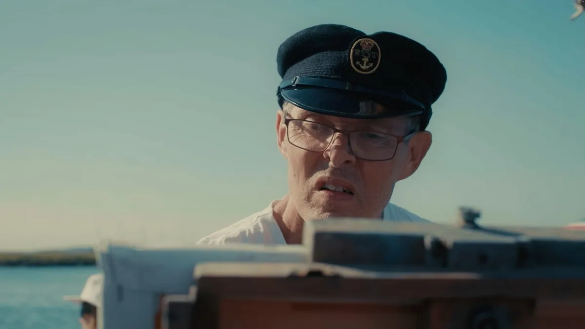 Close-up of a man in glasses and a dark peaked cap with an anchor badge, looking down while painting on a boat, sky and water behind him.
