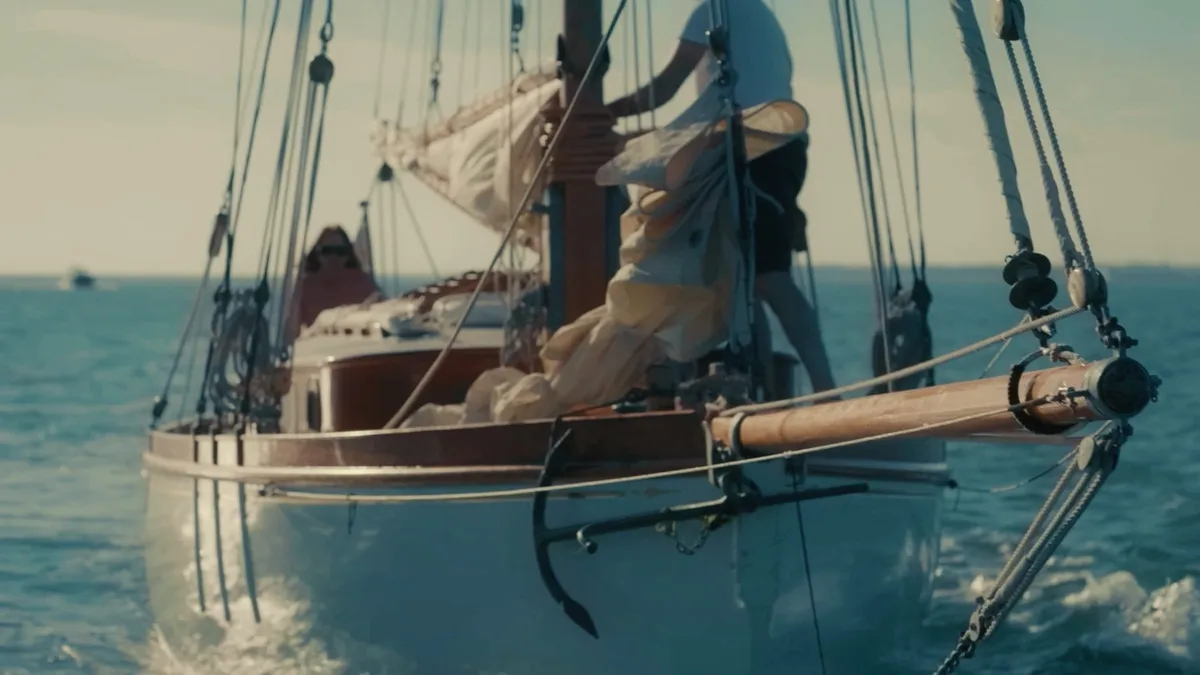 A man in a white T-shirt and dark shorts stands at the mast gathering sailcloth aboard a wooden yacht, anchor visible at the bow, open sea behind.