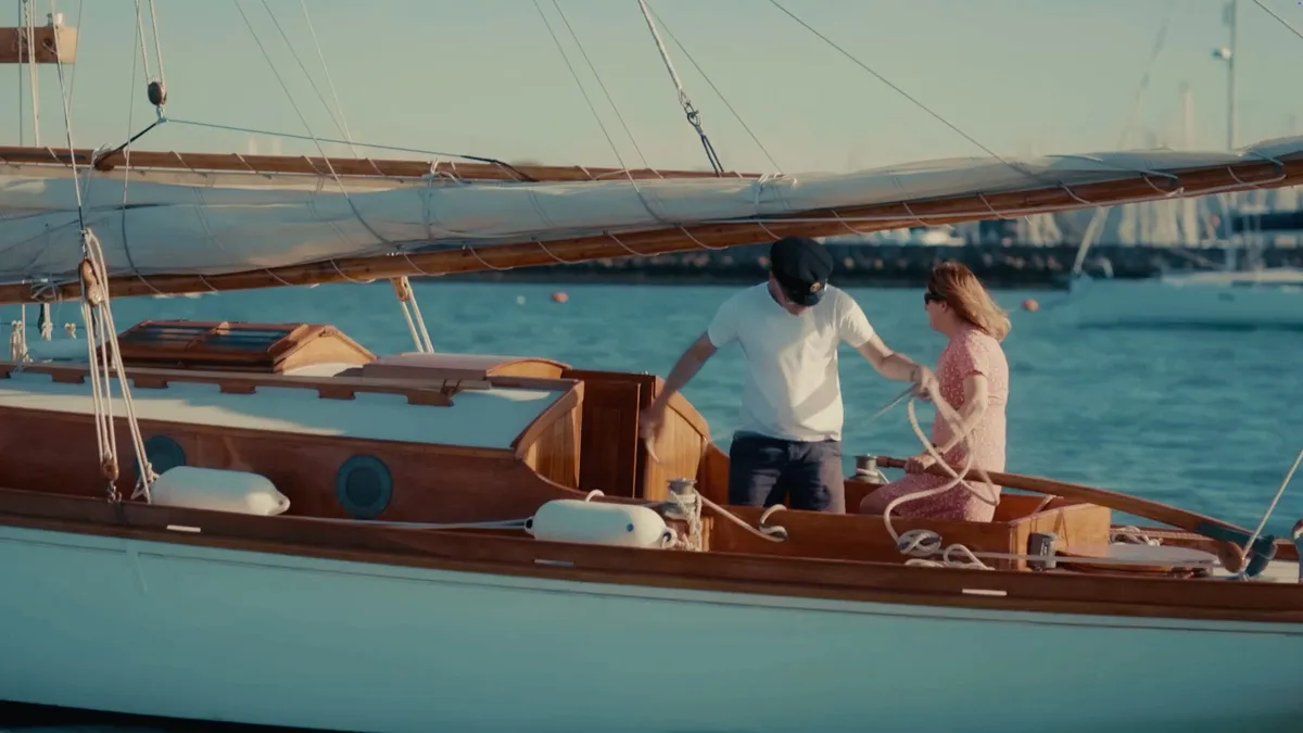 A man in a peaked cap and white T-shirt hauls rope on a wooden sailing boat as a woman in a pink dress helps beside him, marina and moored boats behind.