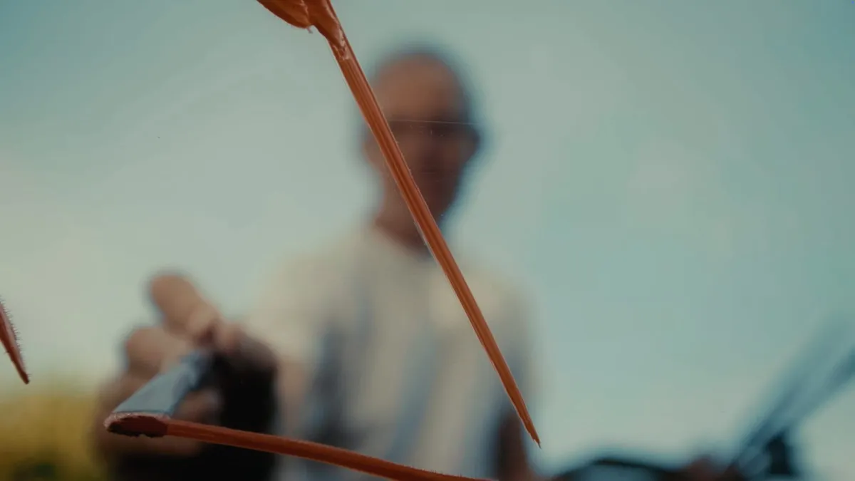 Close-up of a paint-laden brush held up against a blurred grey-haired man in glasses and a pale T-shirt in the background.