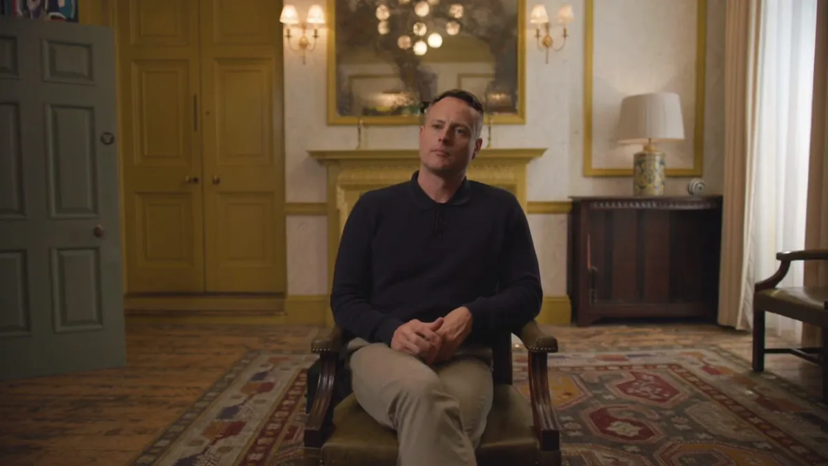 A short-haired man in a navy jumper and beige trousers sits in an armchair, facing camera, in a grand room with yellow panelling.