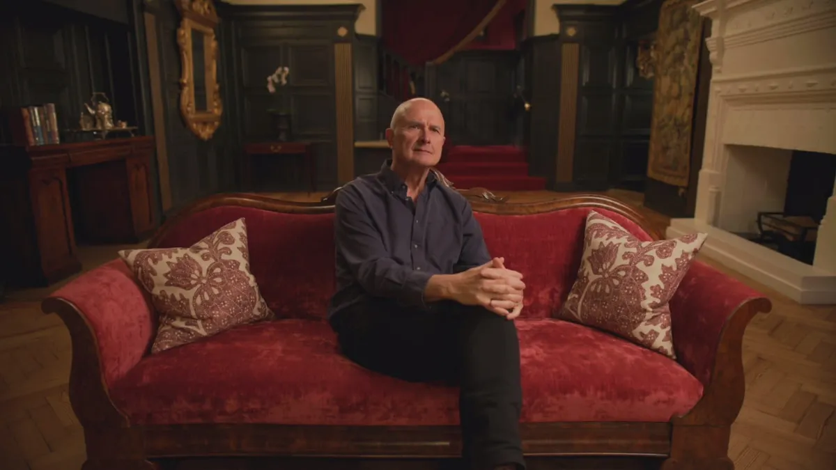A bald man in a dark grey shirt sits on a red velvet sofa, hands clasped, facing camera in a wood-panelled room with a white fireplace.