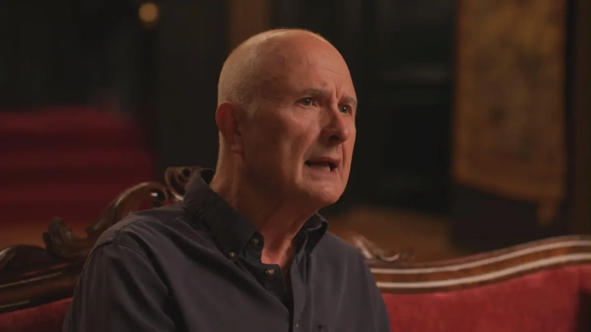 Close-up of a bald man in a dark grey shirt, speaking and looking off-camera, seated on a red velvet sofa in a wood-panelled room.