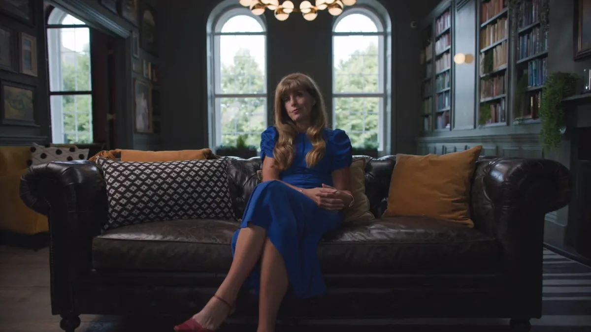 A woman with long wavy blonde hair, in a blue dress, sits on a leather sofa facing camera in a library-style room with arched windows.