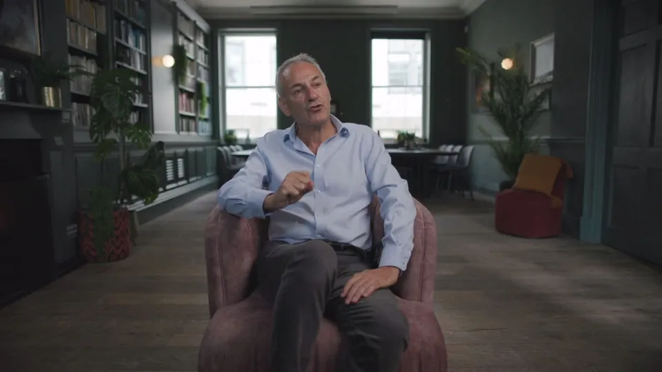 A grey-haired man in a light blue shirt sits in an armchair, gesturing with one hand, facing camera in a panelled library with bookshelves.