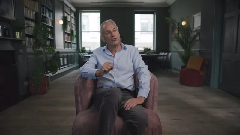 A grey-haired man in a light blue shirt sits in an armchair, gesturing with one hand, facing camera in a panelled library with bookshelves.