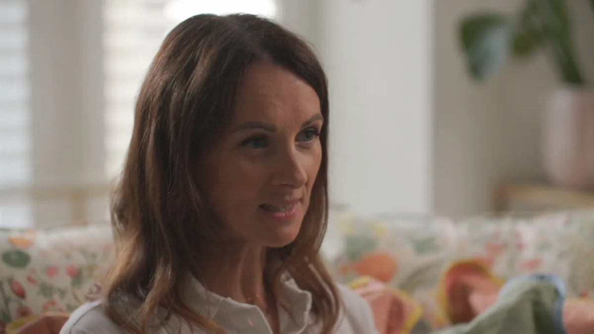 Close-up of a woman with long reddish-brown hair, in a white shirt, looking off-camera on a floral sofa with shuttered windows behind her.
