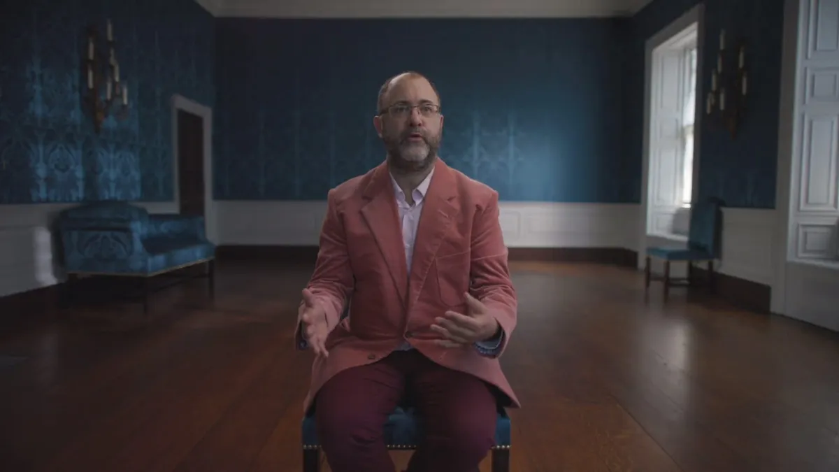 A bald, bearded man in glasses and a pink jacket sits on a chair, gesturing with both hands, facing camera in a blue-panelled room.