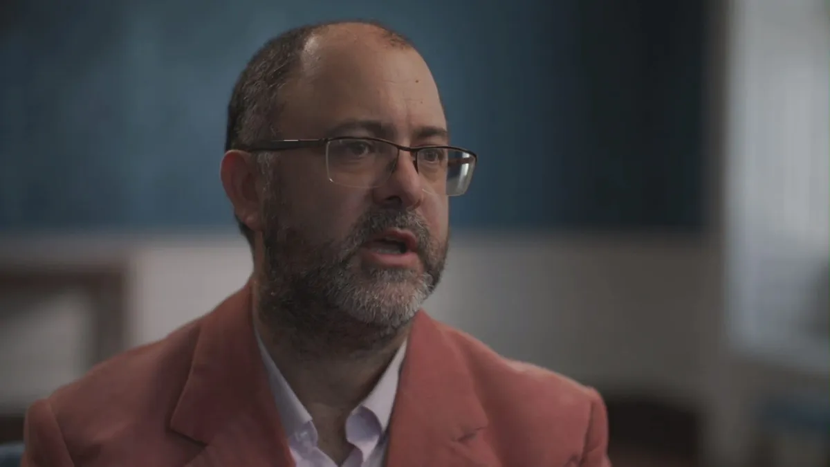 Close-up of a bald, bearded man in glasses and a pink jacket, speaking and looking off-camera, with a blue-panelled room behind him.