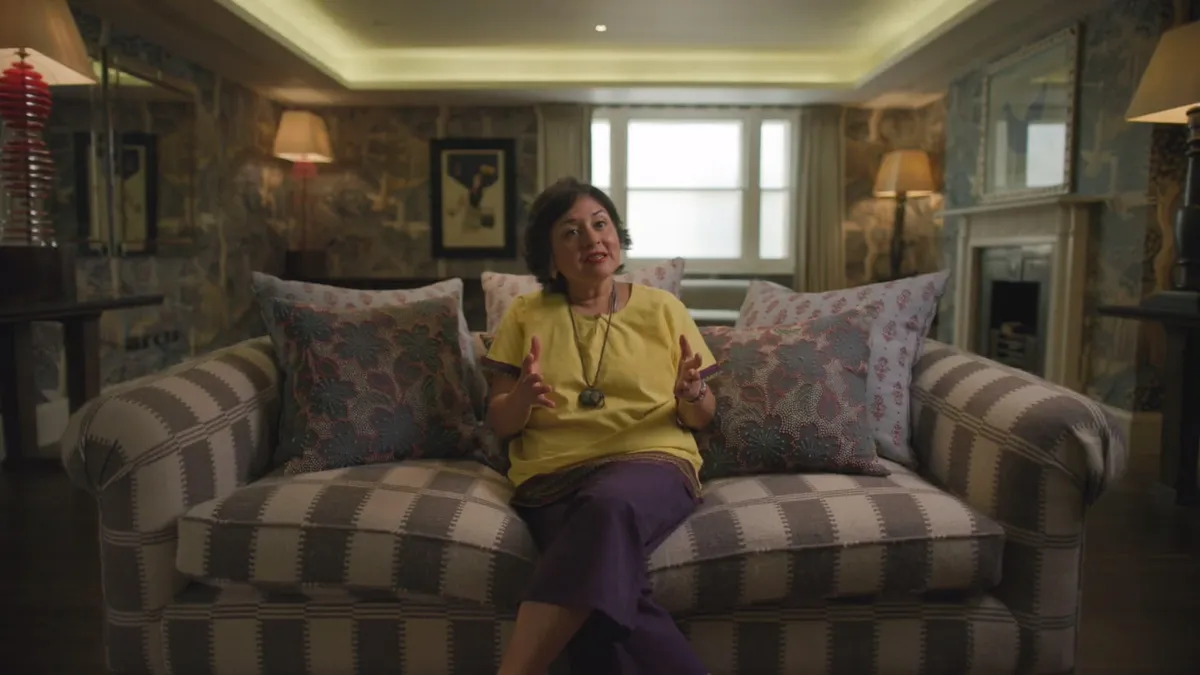 A short-haired woman in a yellow top and purple skirt sits on a checked sofa, gesturing with both hands, facing camera in a patterned sitting room.