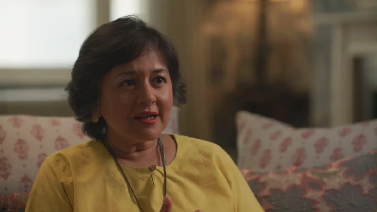 Close-up of a short-haired woman in a yellow top, speaking and looking off-camera, on a patterned sofa with cushions behind her.