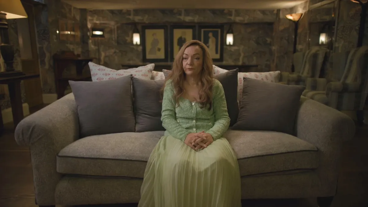 A woman with long wavy reddish hair, in a green top and pale green skirt, sits on a grey sofa, hands clasped, facing camera in a patterned room with framed portraits.