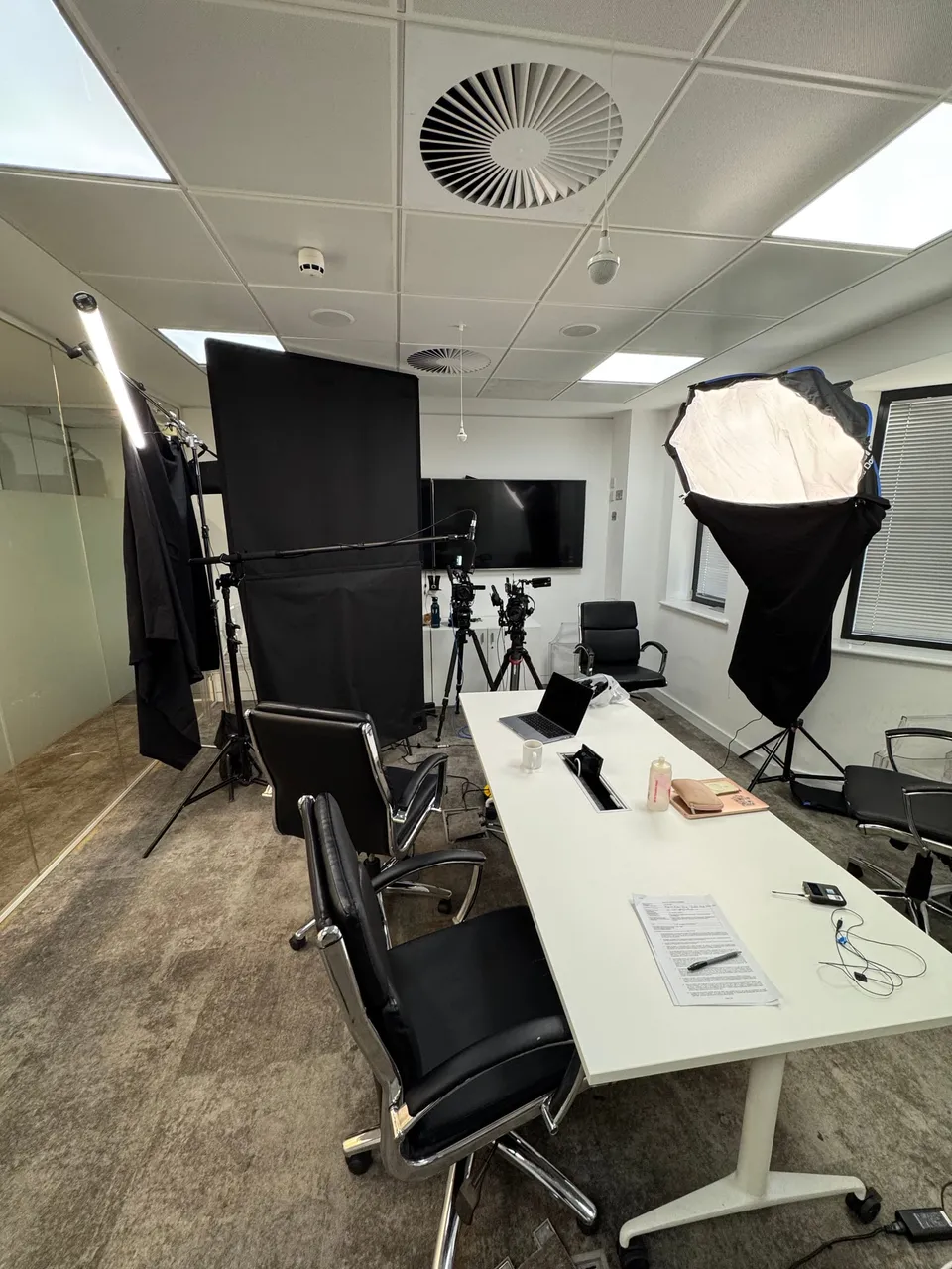 An empty office set up for filming, with a white table, black office chairs, camera tripods, a softbox light and backdrop, and a wall-mounted screen.