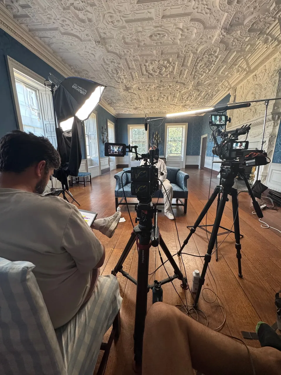 Camera crew filming an interview in an ornately plastered blue-panelled room, with tripod-mounted cameras, monitors and a softbox light facing a blue sofa.
