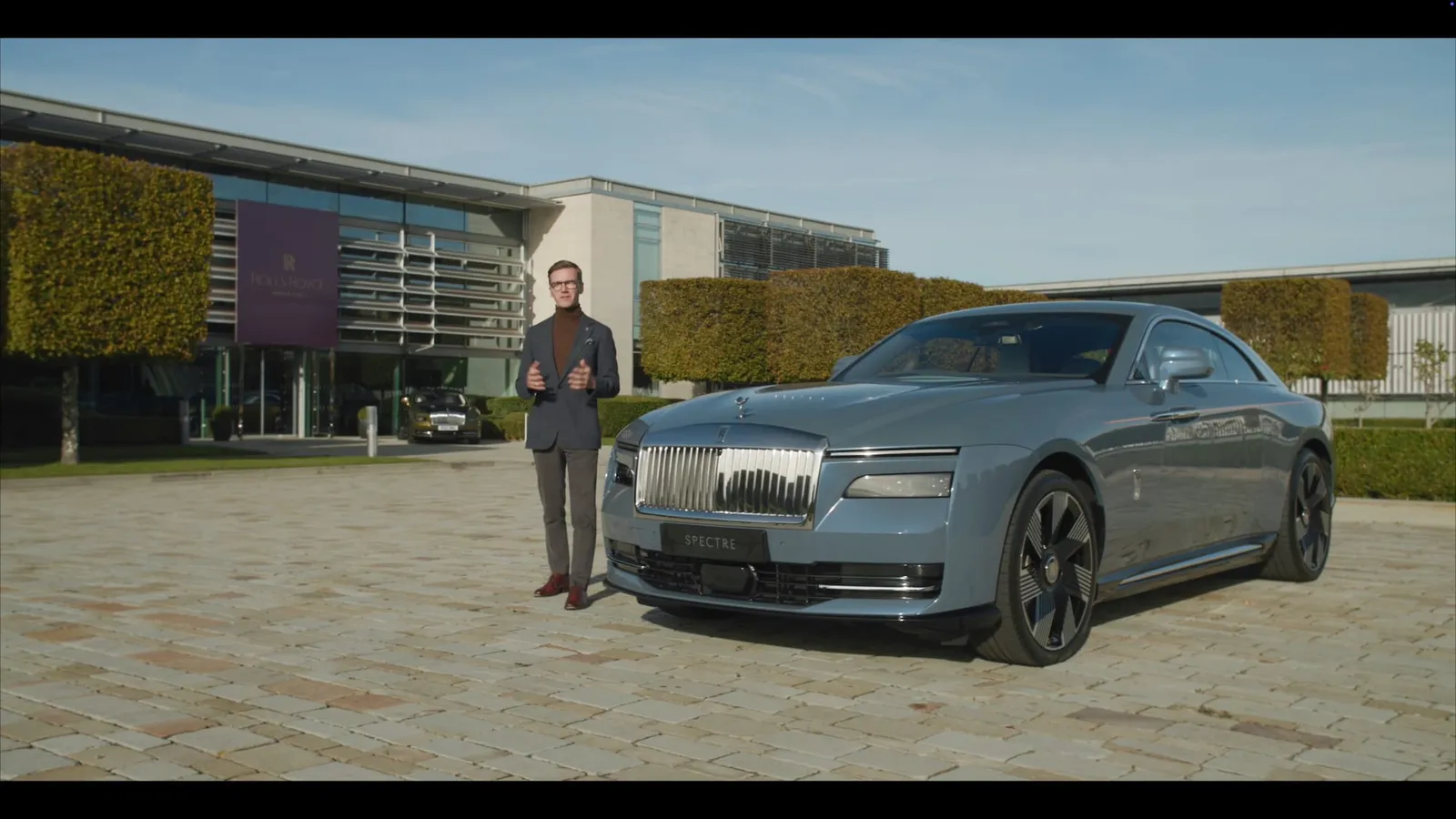 A short-haired man in a grey suit stands beside a grey saloon car labelled "Spectre", facing camera outside a modern building with hedges.