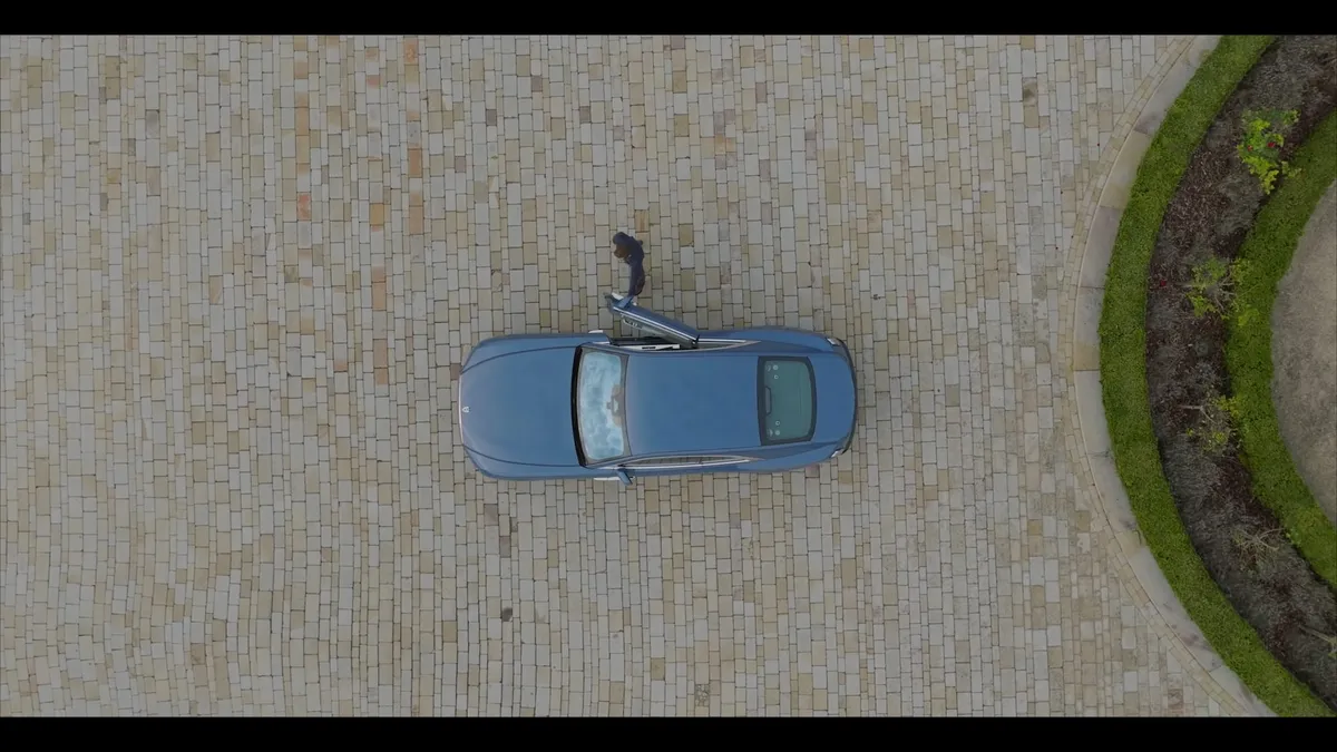 Aerial view of a blue car on a cobbled driveway, with a person standing beside the open bonnet and a curved hedge to the right.