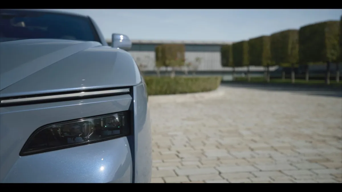 Close-up of a silver car's headlight and bonnet, with a paved driveway, hedges and trees blurred in the background.