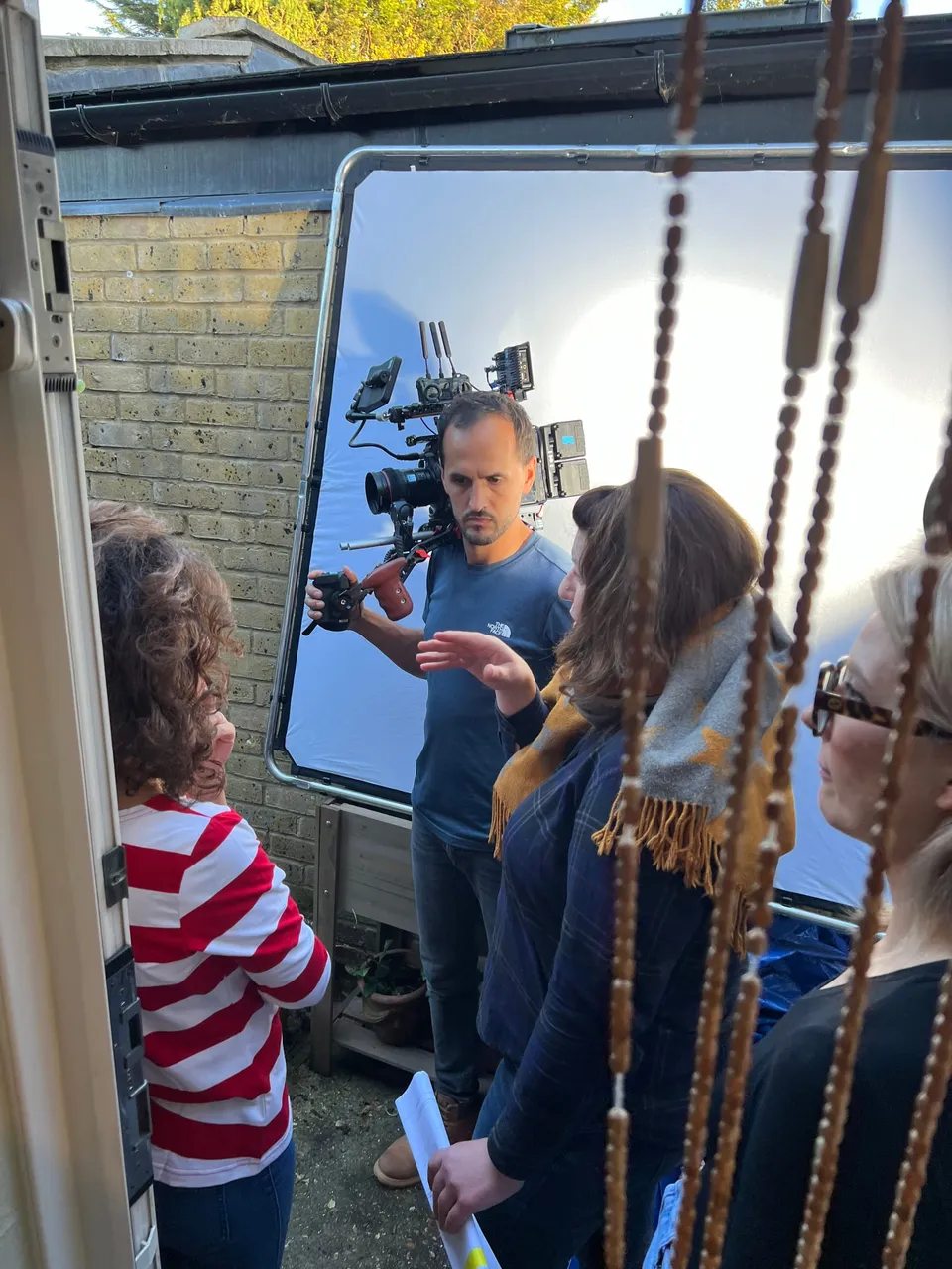 A camera operator in a blue T-shirt holds a camera rig behind a large white reflector, watched by three crew members through a beaded curtain.