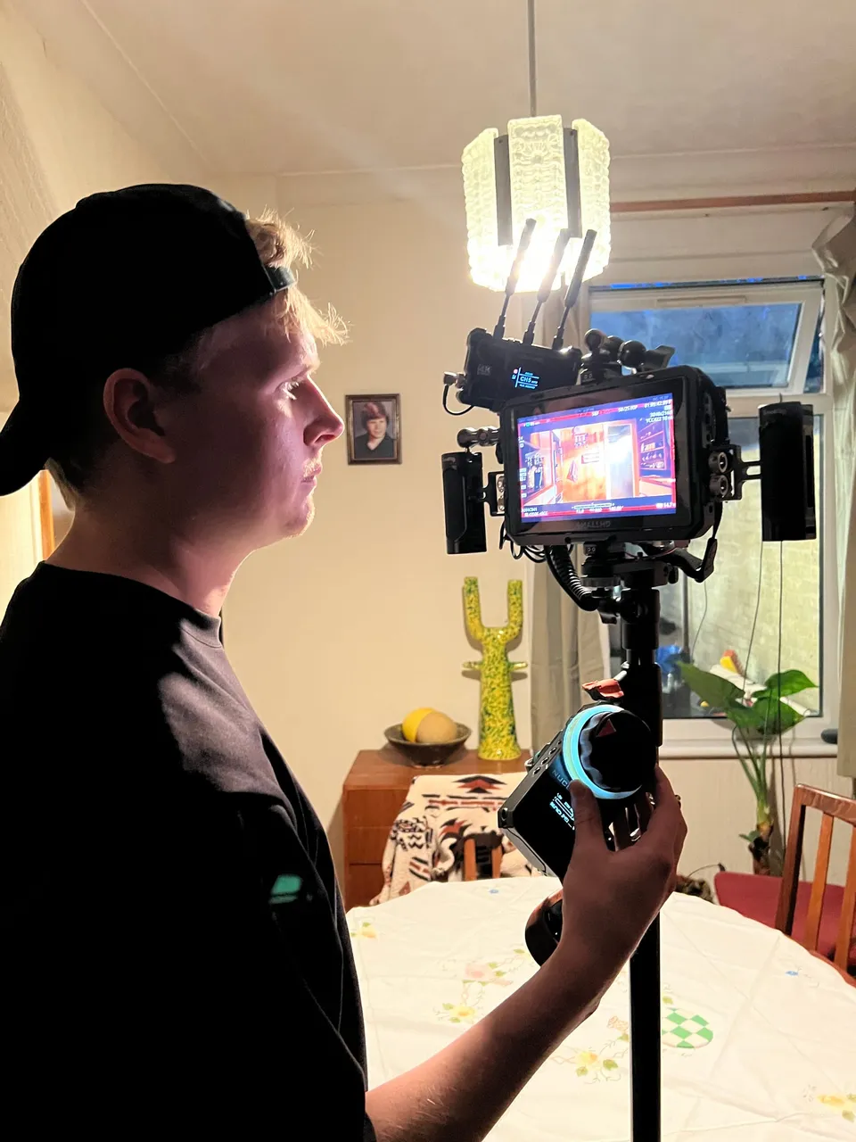 In profile, a man in a black cap and dark T-shirt operates a focus monitor rig at a domestic dining table lit by a pendant lamp.