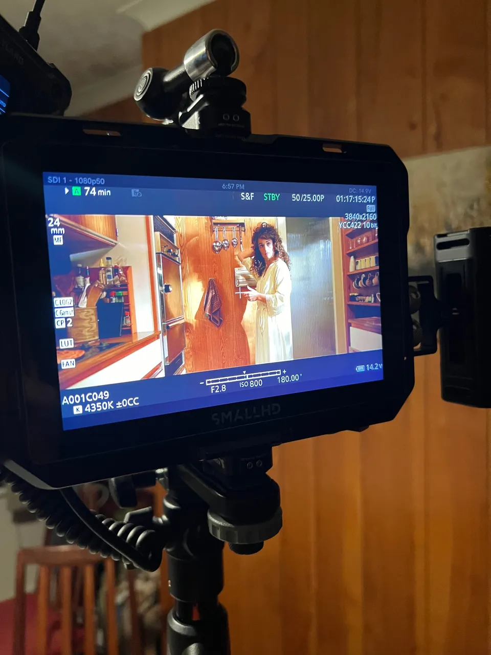 A camera monitor mounted on a tripod shows a curly-haired woman in a cream robe standing in a wood-panelled kitchen doorway.