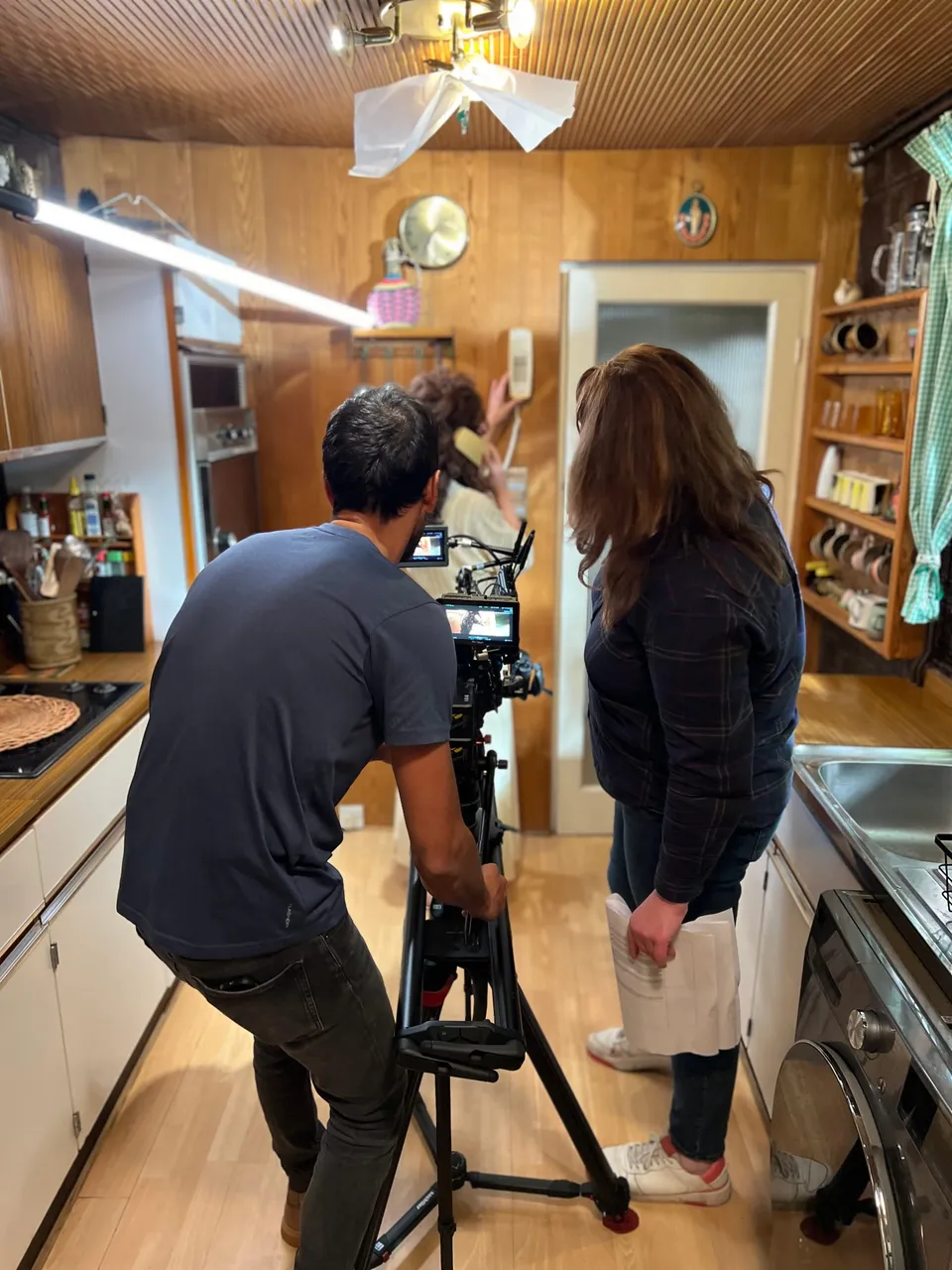A camera operator in a blue T-shirt films on a slider rig in a wood-panelled kitchen, beside a dark-haired woman in navy holding fabric.