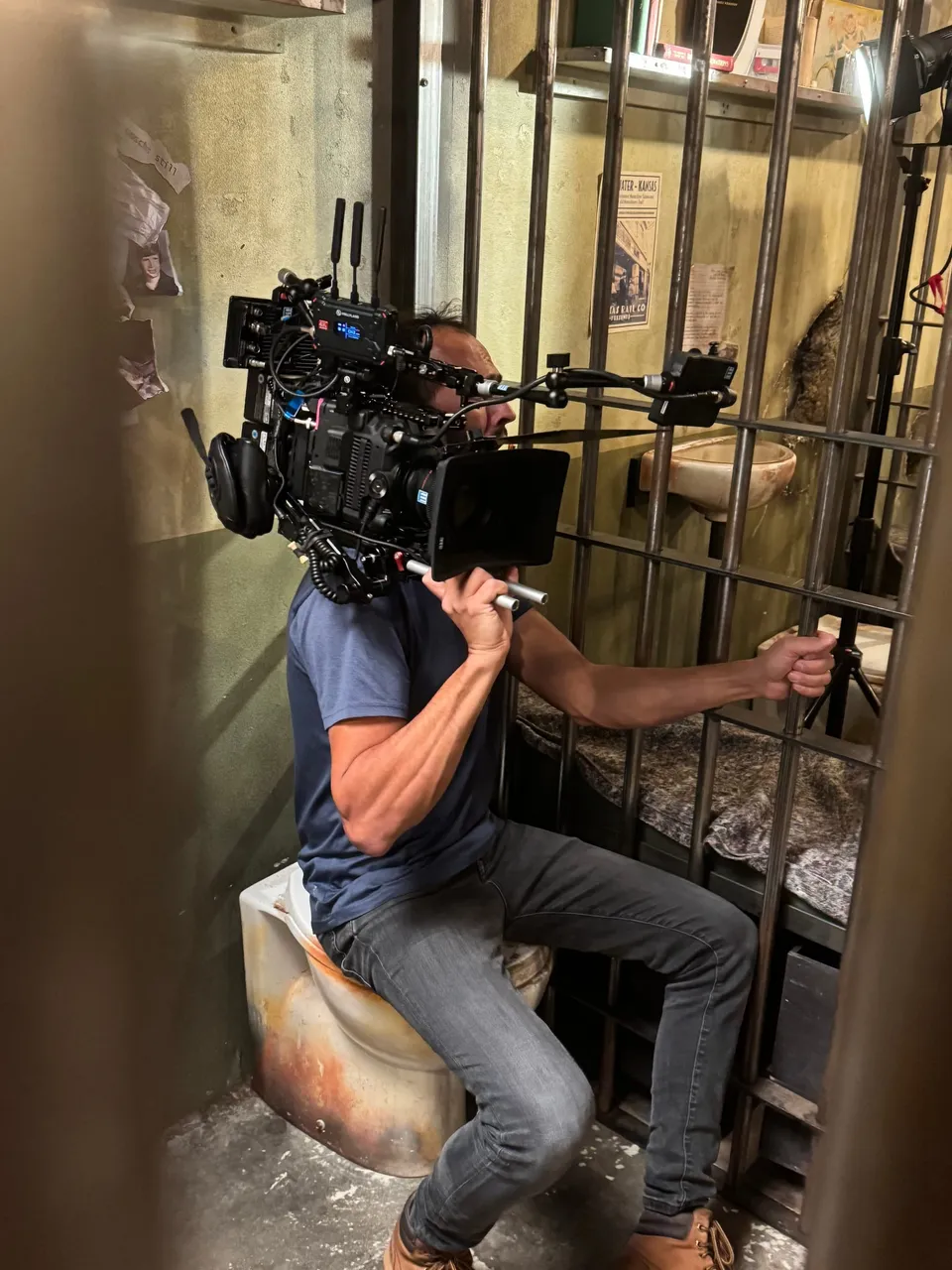 A camera operator in a blue T-shirt sits perched on a prop toilet, filming through jail bars in a cell set with a bunk and posters on the wall.