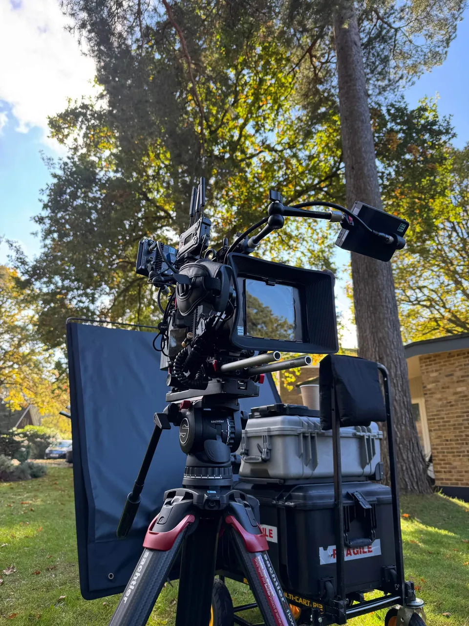A camera rig on a tripod with a monitor and equipment cases, set up outdoors on grass in front of trees and a brick building.