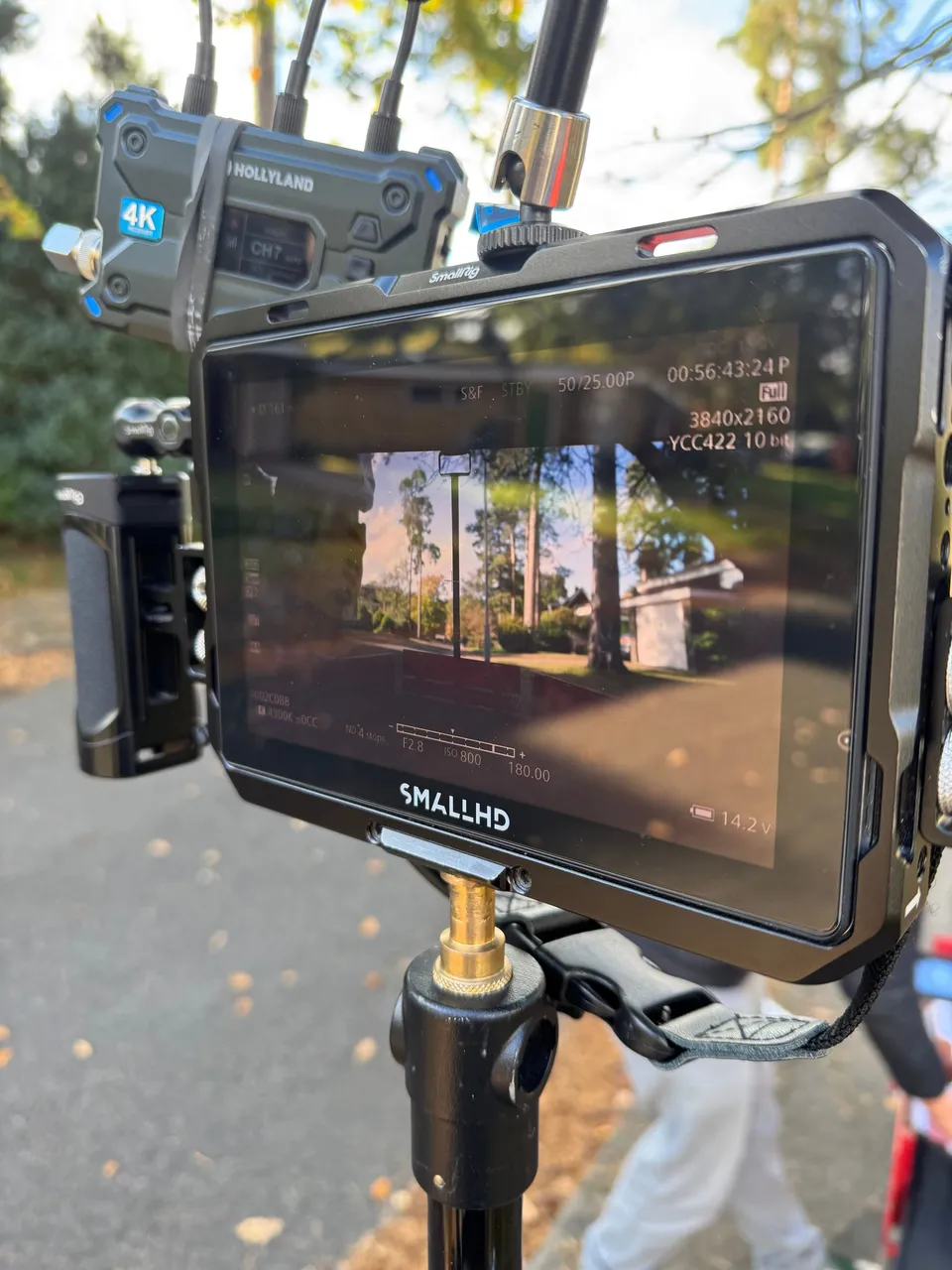 A camera monitor on a tripod displays a tree-lined street scene, with a wireless receiver mounted above it outdoors.