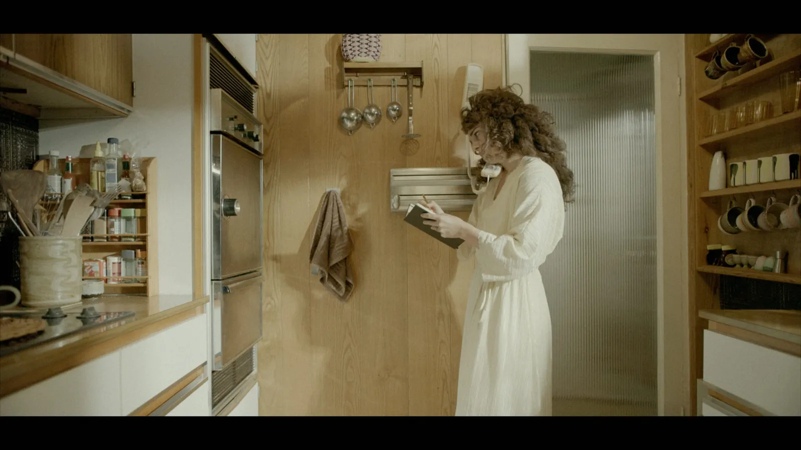 A curly-haired woman in a cream robe stands by a wall-mounted phone in a wood-panelled kitchen, writing in a notebook.