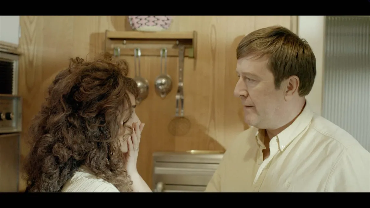 A curly-haired woman touches her face, facing a man with brown hair in a cream shirt, in a wood-panelled kitchen with hanging utensils.