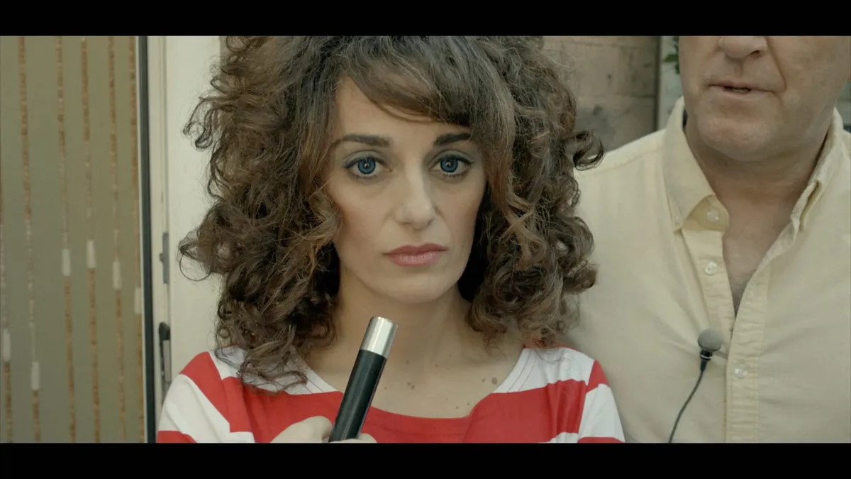 Close-up of a curly-haired woman in a red-and-white striped top holding a microphone stand, facing camera, a man in a cream shirt behind her.