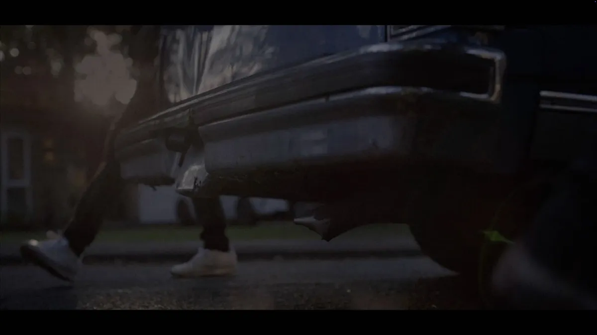 Low-angle close-up of a vintage car's rear bumper, with a person in dark trousers and white trainers running past on a wet street.