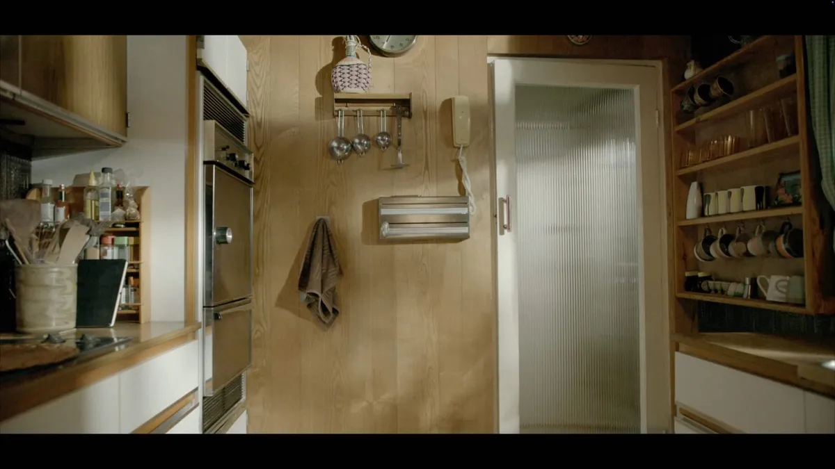 A wood-panelled kitchen wall with a corded telephone, hanging measuring cups, a towel and a paper towel dispenser, beside a frosted-glass door.