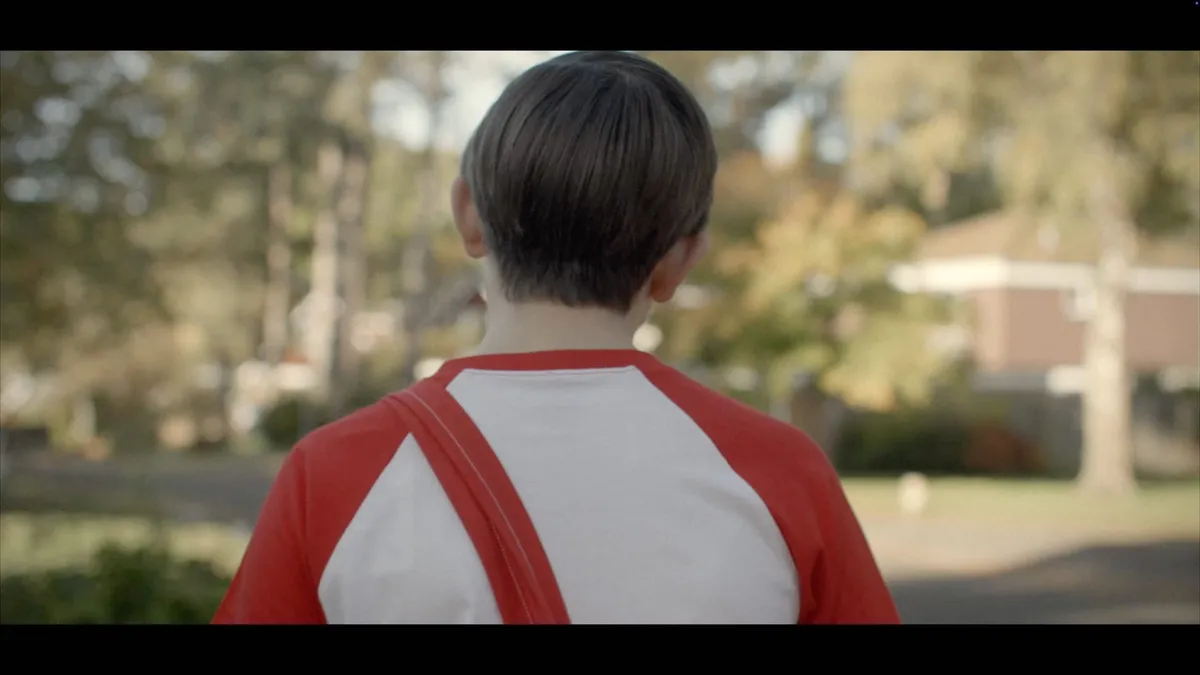 A boy with neat brown hair, in a red and white raglan T-shirt, seen from behind, facing away on a suburban street with trees and a house blurred beyond.