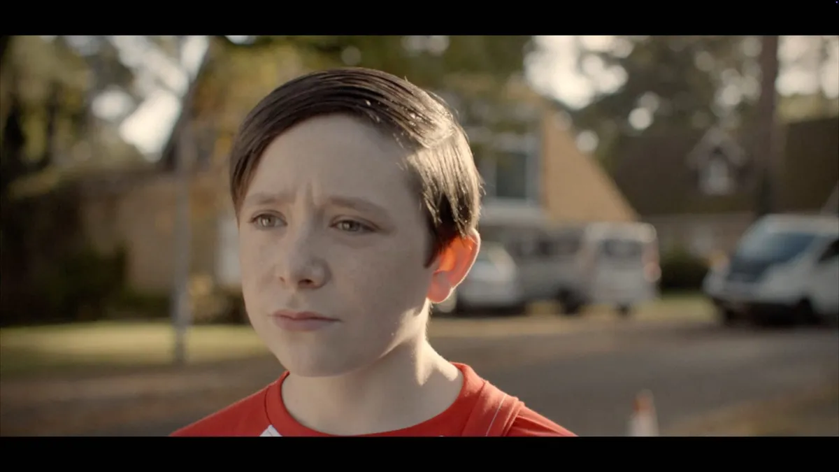 Close-up of a boy with neat brown hair, in a red top, looking off-camera with a concerned expression on a suburban street.