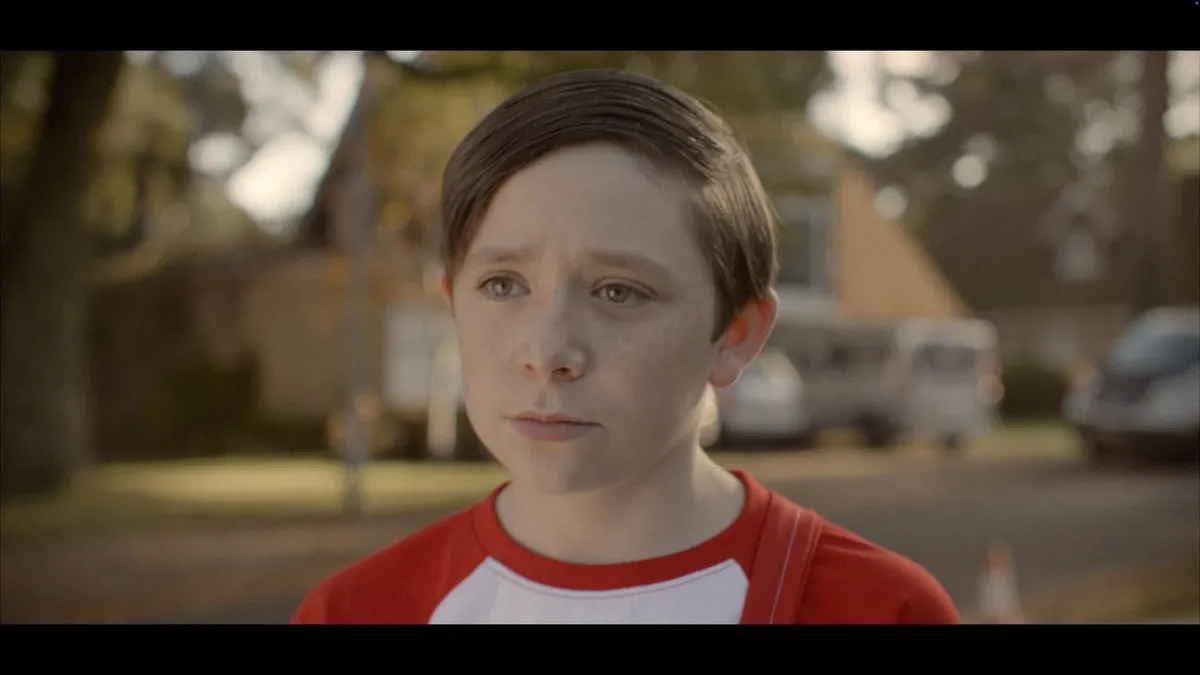 Close-up of a boy with neat brown hair, in a red and white raglan T-shirt, looking off-camera with a concerned expression, houses blurred behind him.